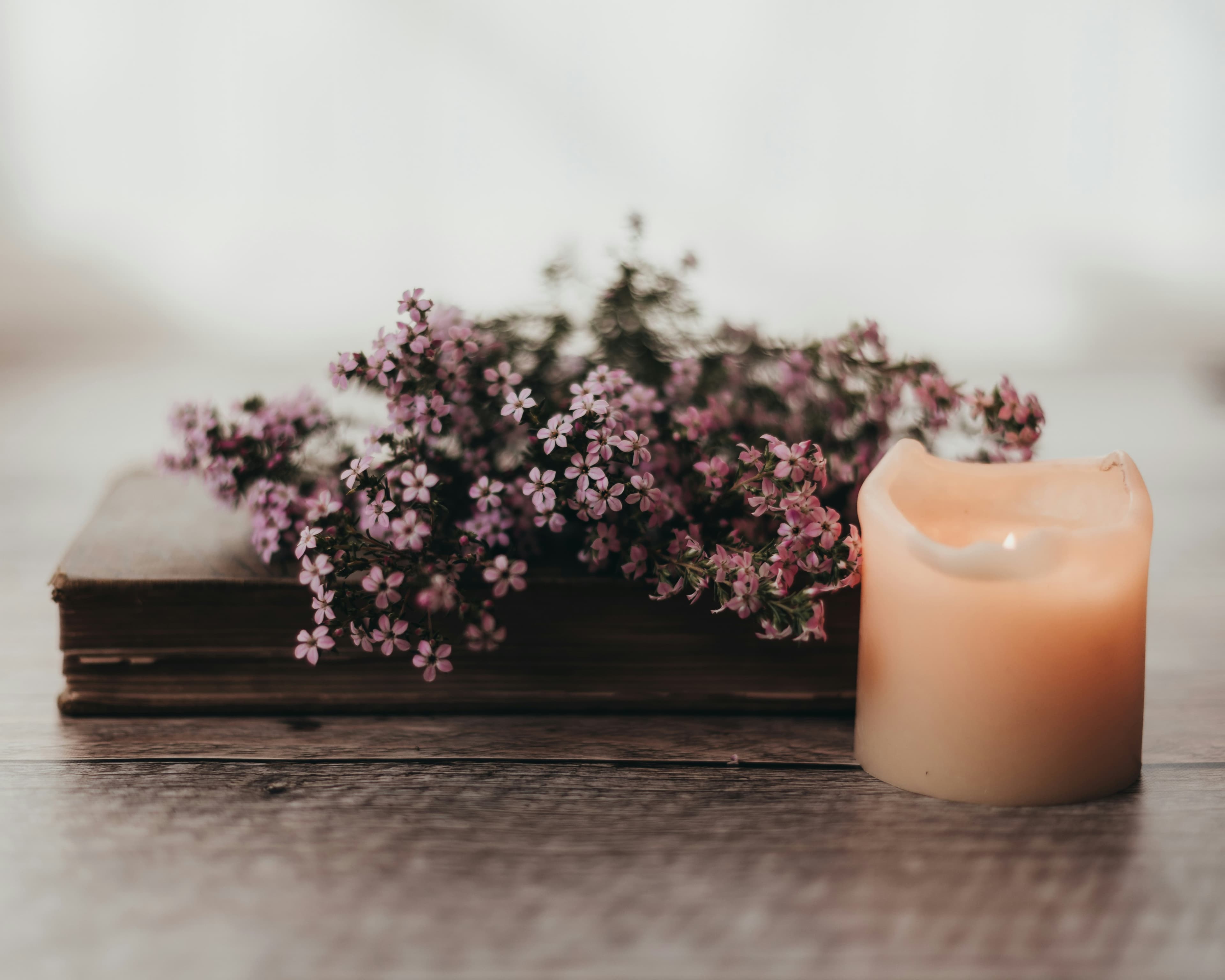 A candle beside a worn book topped with small pink flowers on a wooden table in soft natural light.